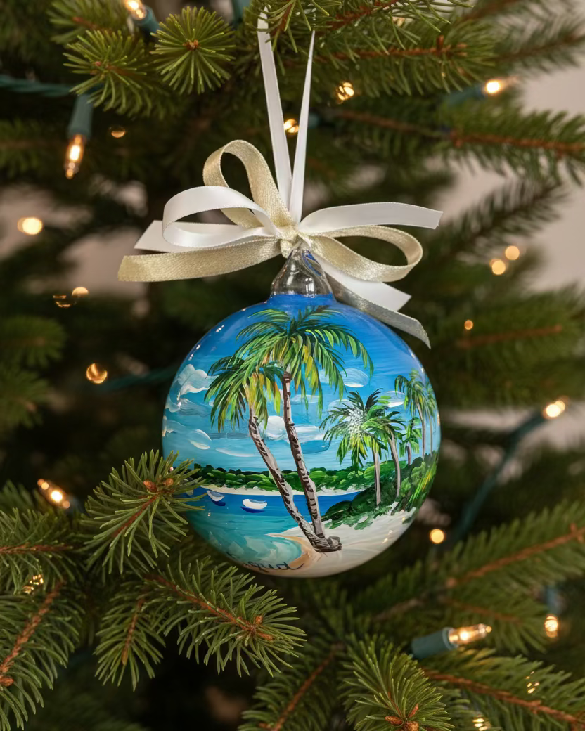 Hand-painted glass Antigua ornament hanging on a Christmas tree, decorated with palm trees, a tropical beach, turquoise sea, and a gold-and-white ribbon.