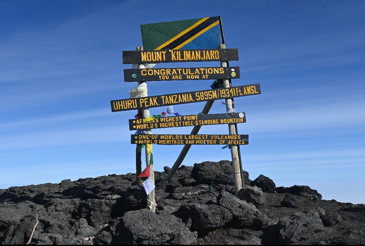 Mount Kilimanjaro Uhuru Peak summit sign "Congratulations you are now at Uhuru Peak Tanzania 5895m" with Tanzania flag, black volcanic rocks and blue sky, Africa highest point
