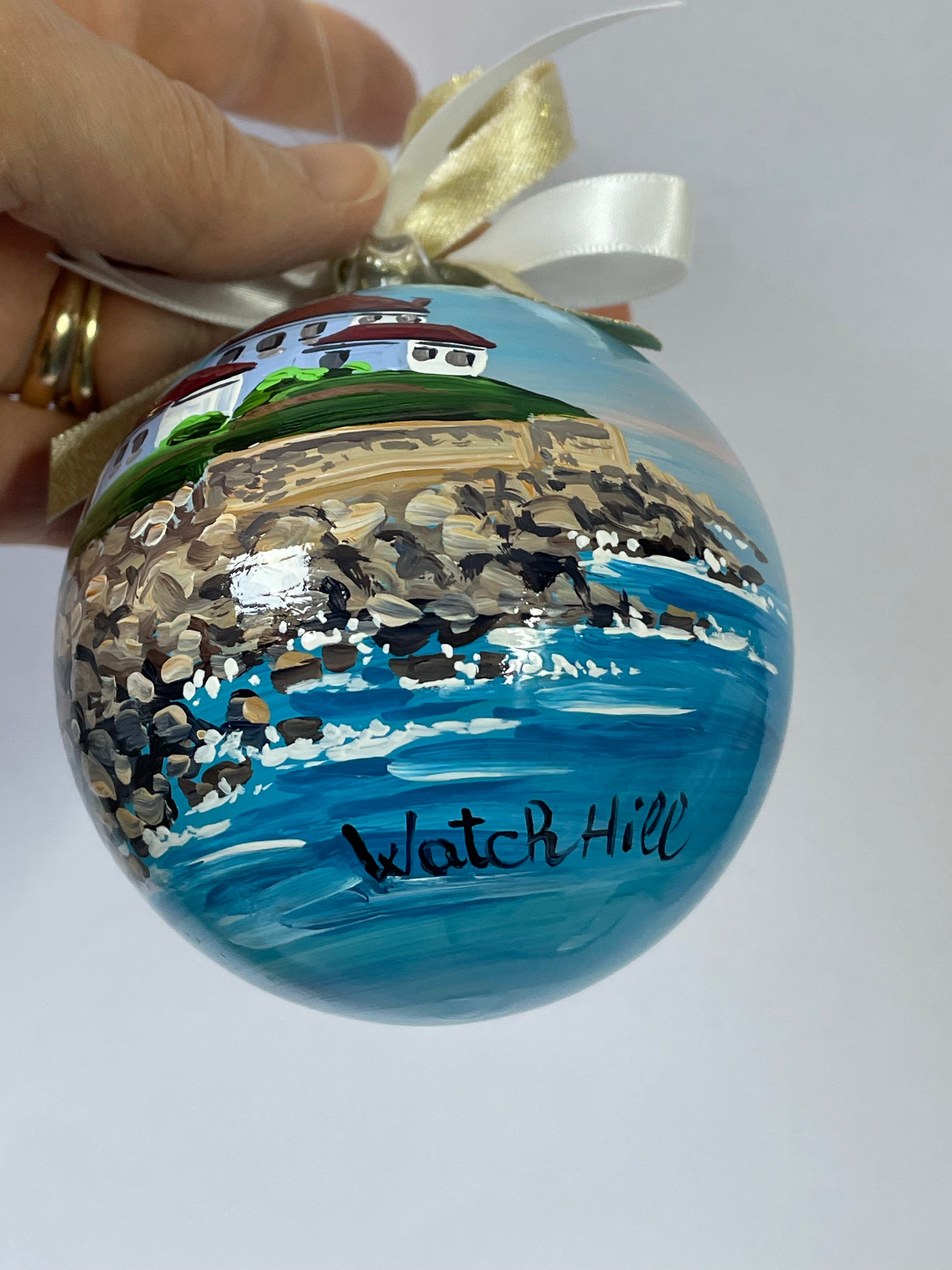 Hand holding hand-painted glass ornament – Watch Hill inscription with lighthouse, rocky shore and blue waves, Rhode Island
