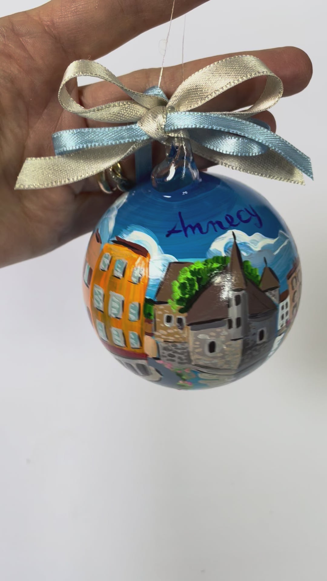 Hand-painted glass ornament – Annecy inscription with Palais de l'Isle stone tower, colourful buildings, snow-capped mountains and turquoise canal, blue and gold ribbon, France souvenir